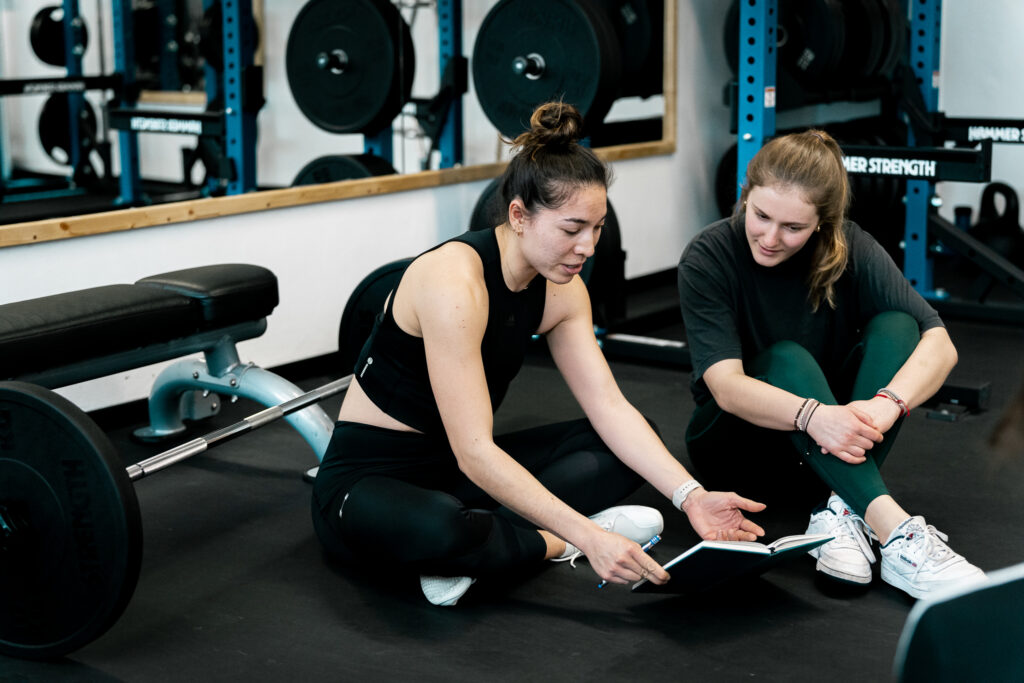 Personal Trainerin Ellise Meyer blickt mit einer Kundin zusammen in ein Notizbuch.