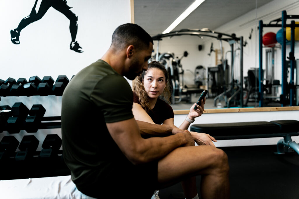 Personal Trainerin Ellise Meyer hockt neben einem Kunden, der auf einer Langhantelbank sitzt, und erklärt ihm etwas.