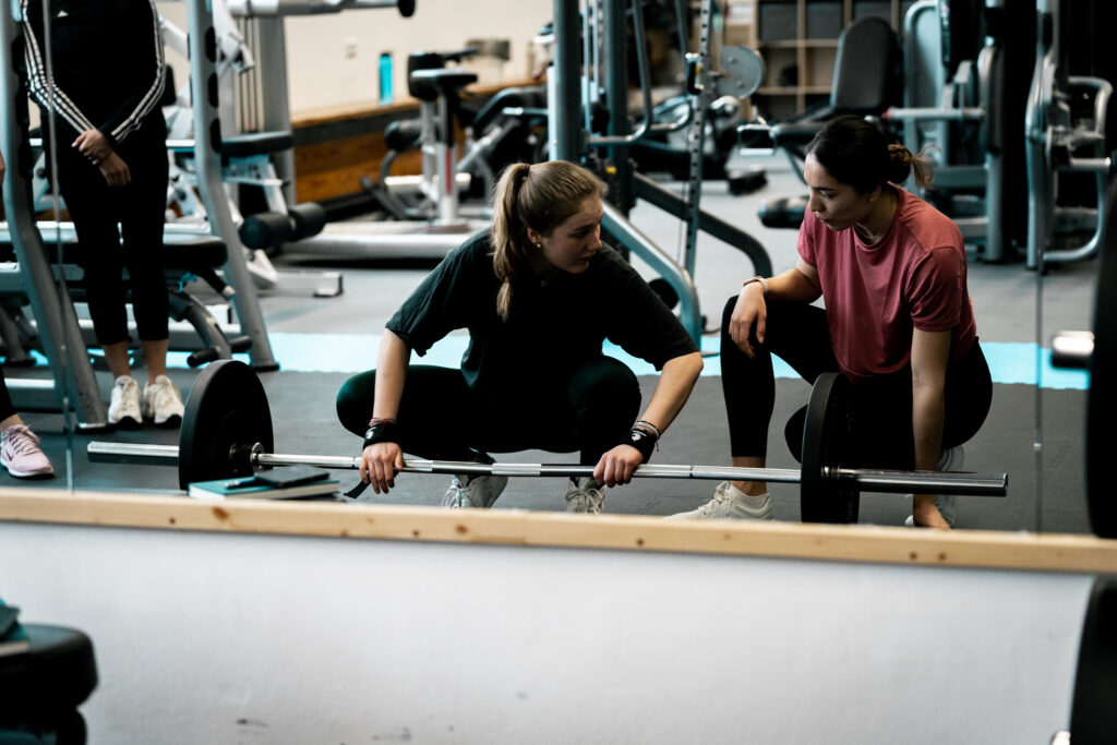 Personal Trainerin Ellise Meyer sitzt mit einer Kundin vor einer Langhantel.